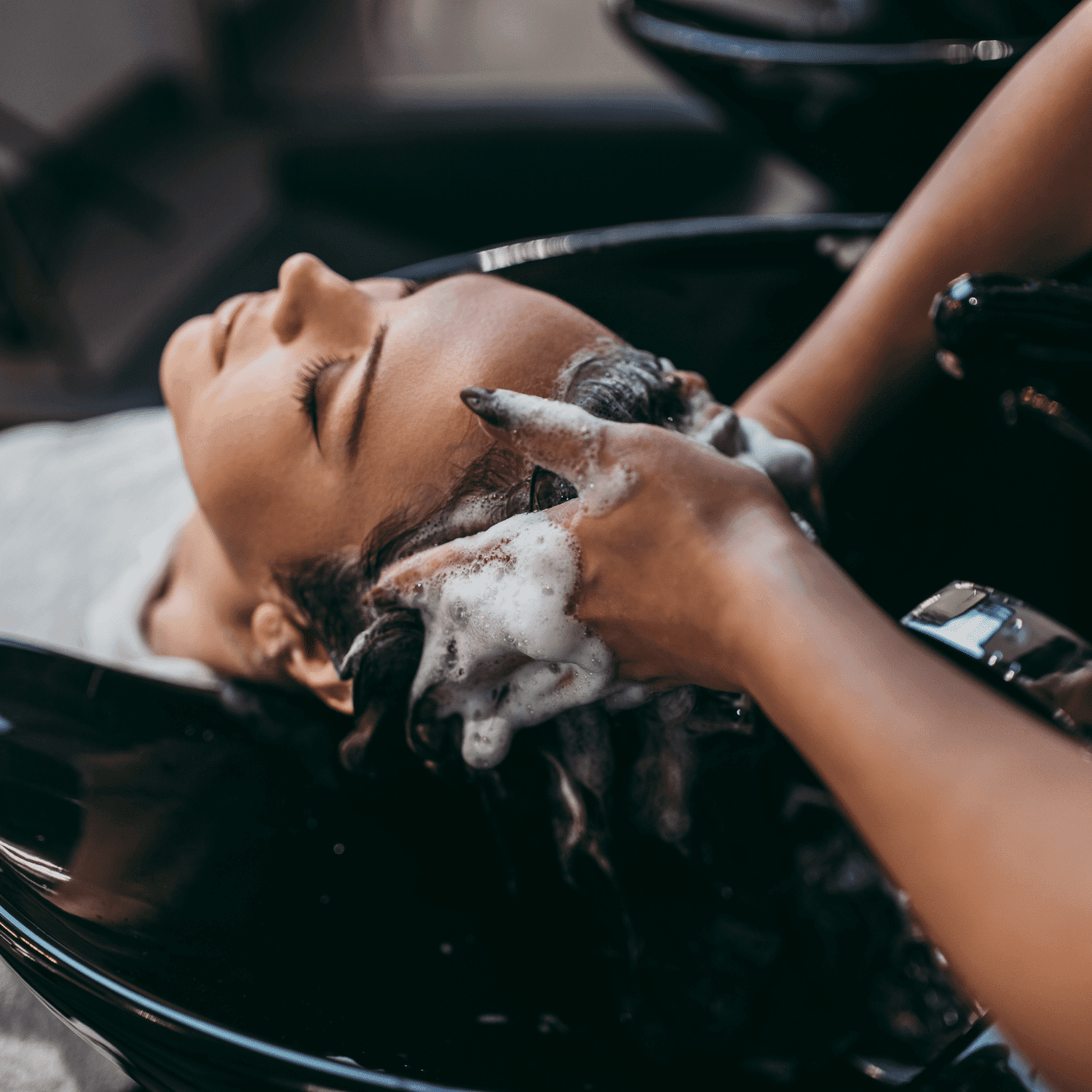 Person getting hair washed at a salon, head tilted back, and hands applying shampoo.